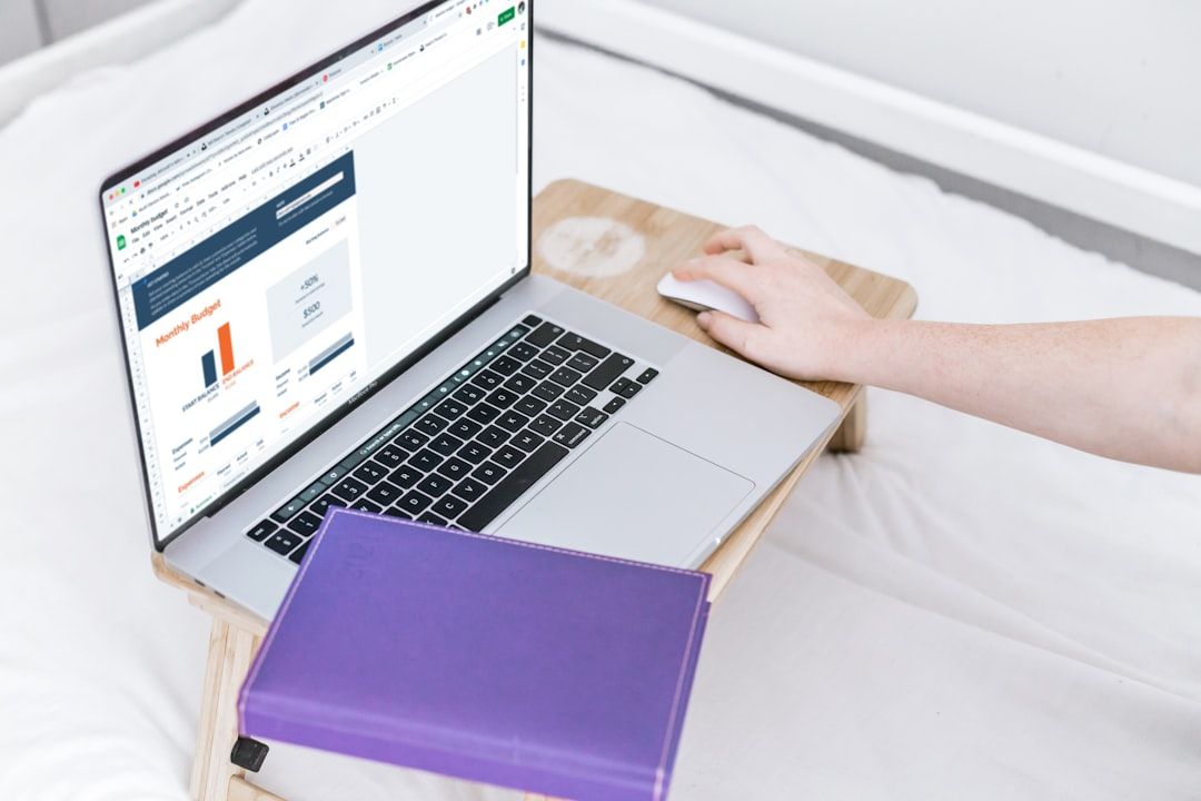Eine Person sitzt auf einem Bett und arbeitet mit einem Laptop. Auf dem Bildschirm wird eine Tabelle angezeigt. Auf dem Schoßtisch neben dem Laptop liegt ein geschlossenes, violettes Notizbuch aus Aachen.