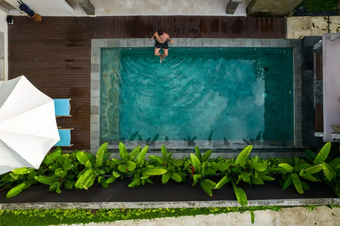 Eine Person schwimmt in einem rechteckigen Pool, der von Grünflächen, Holzterrassen und einer Terrasse mit einem weißen Sonnenschirm und blauen Liegestühlen umgeben ist.