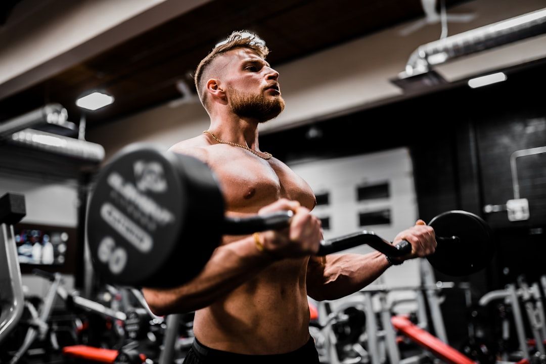 Ein muskulöser Mann macht in einem Fitnessstudio in Aachen Bizepscurls mit einer Langhantel. Er ist mit nacktem Oberkörper und konzentriert, umgeben von Fitnessgeräten.