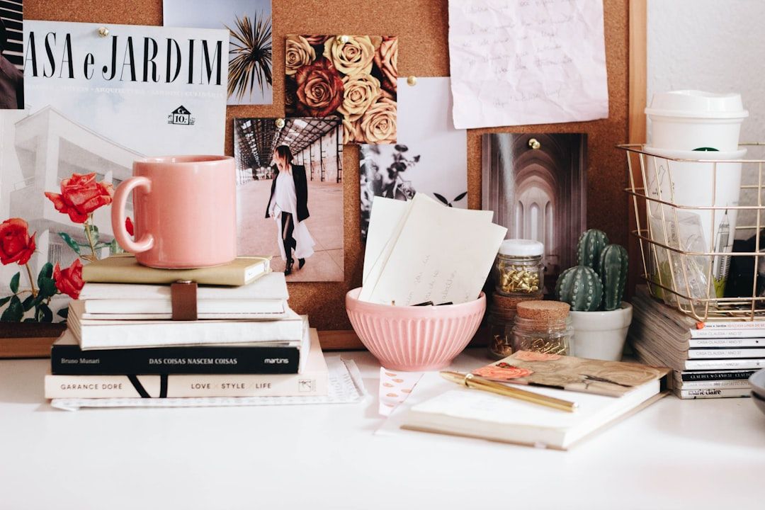 Ein unordentlicher Schreibtisch mit Büchern, einer rosa Tasse, einer Schale mit Papieren, einem Kaktus und einer Korktafel, die mit verschiedenen Bildern aus Aachen geschmückt ist, wird ergänzt durch einen Drahtkorb, in dem ein Pappbecher hängt.