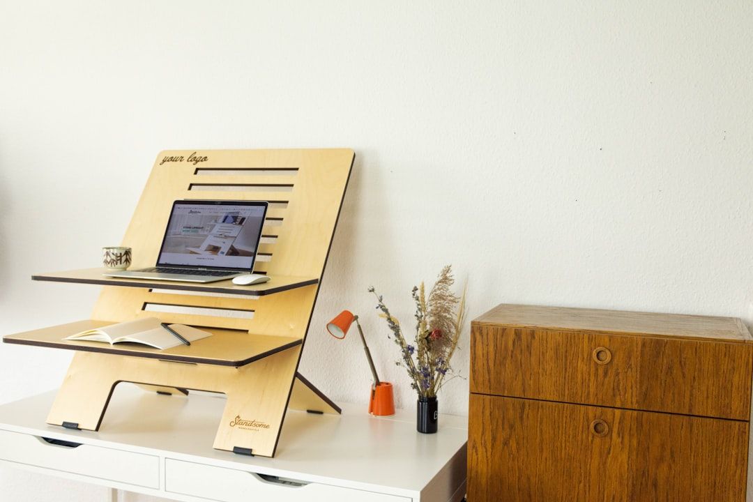 Ein Stehpult aus Holz mit Laptop, Notizbuch und Kaffeetasse steht auf einem weißen Tisch und erinnert an die charmanten Innenräume Aachens. Eine kleine Lampe und getrocknete Blumen in einer Vase vervollständigen die Szene. Daneben steht ein Holzschrank.