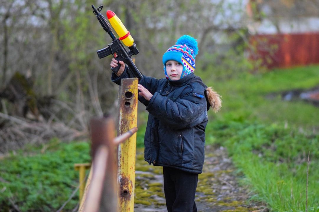 Ein Kind mit bunter Wintermütze und dunklem Mantel steht im Freien in Aachen und hält eine Wasserpistole in der Hand. Im Hintergrund sind Grünflächen und ein Weg zu sehen.