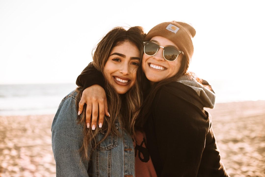 Zwei Frauen stehen an einem Strand in Aachen, lächeln und umarmen sich. Eine trägt eine Jeansjacke, die andere eine Sonnenbrille und eine Beanie. Im Hintergrund geht die Sonne unter.