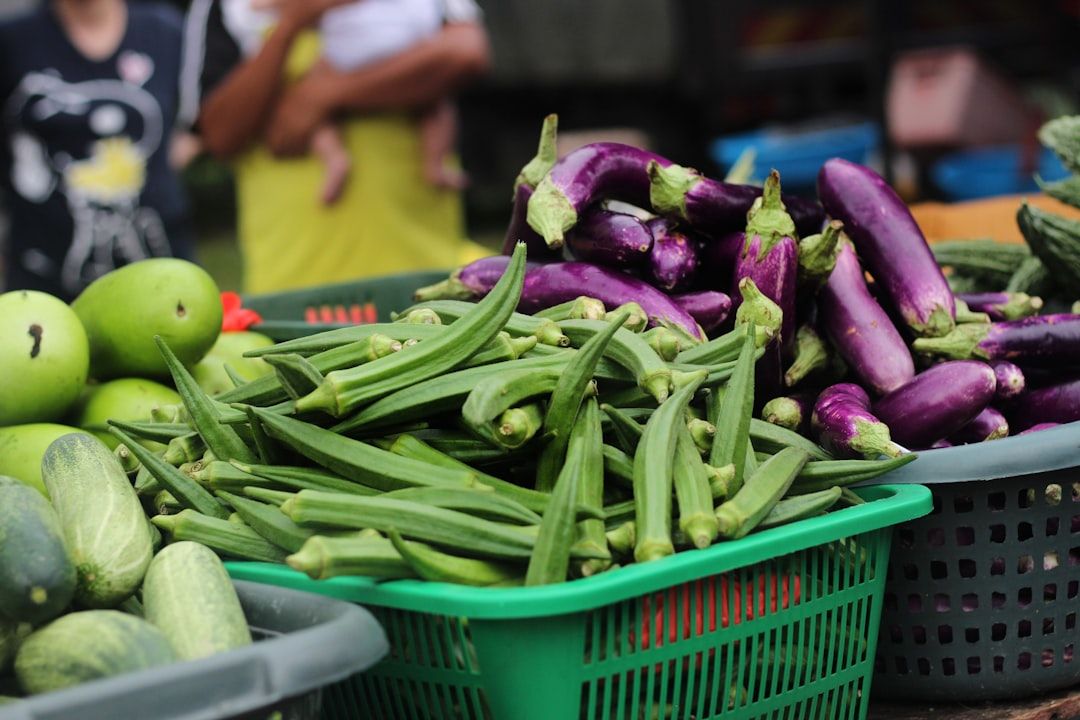 An einem Marktstand in Aachen werden frische grüne Okraschoten, Gurken und violette Auberginen in Plastikkörben angeboten. Im Hintergrund ist eine Person zu sehen, die ein Kind hält.