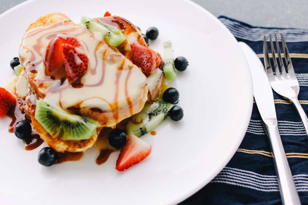 Ein Teller French Toast, belegt mit geschnittenen Erdbeeren, Kiwi, Blaubeeren und beträufelt mit Sirup und Sahne, erinnert an die köstlichen Leckereien, die man in Aachens charmanten Cafés findet, und steht neben einer gefalteten gestreiften Serviette mit Messer und Gabel.