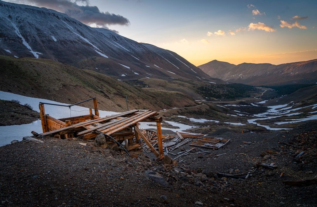 Hölzerne Überreste eines verfallenen Gebäudes in einer Berglandschaft bei Sonnenuntergang, im Hintergrund Schneeflecken und ein an Aachen erinnerndes Tal.
