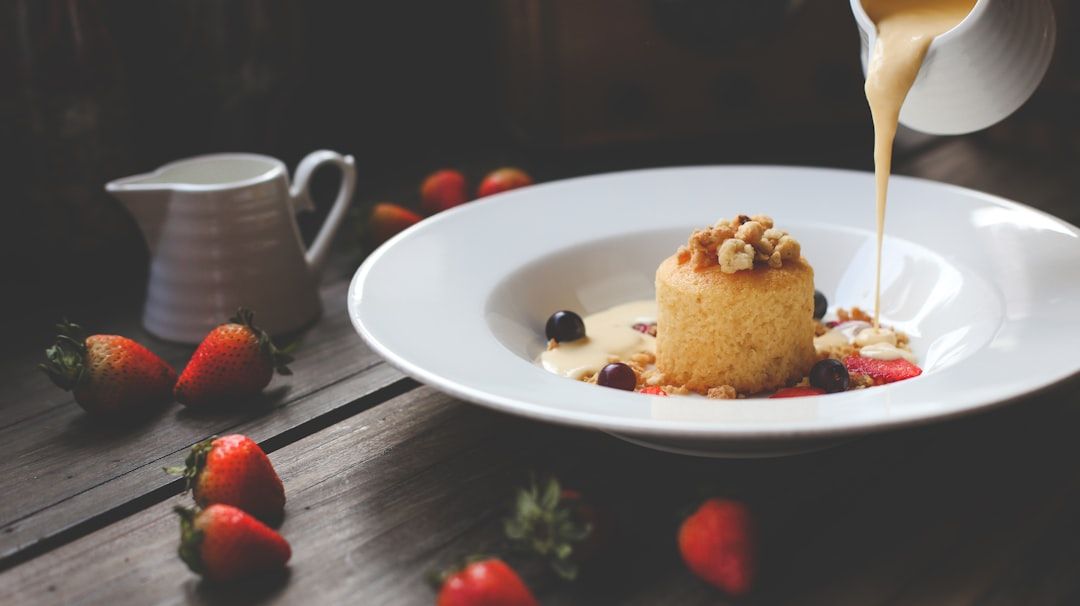 Ein kleiner Kuchen in einer weißen Schüssel wird mit Streuseln belegt und mit einer cremigen Soße übergossen. Auf einer Holzfläche liegen Erdbeeren und Heidelbeeren, die an köstliche Aachener Desserts erinnern.