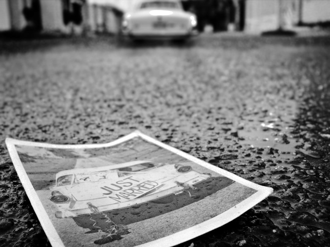 Ein Schwarzweißfoto eines „Just Married“-Schildes auf der Rückseite eines Autos liegt auf einer nassen, strukturierten Oberfläche in Aachen, im Hintergrund ist ein weiteres unscharfes Auto zu sehen.