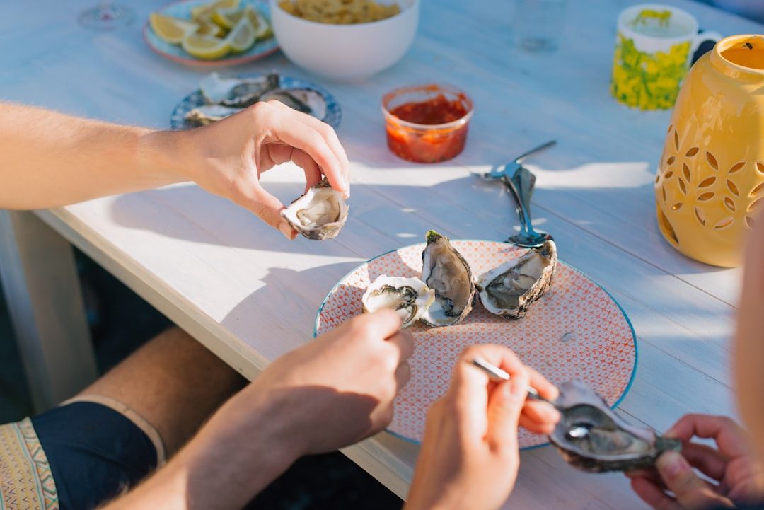 Zwei Personen sitzen an einem Tisch in Aachen, öffnen und essen Austern. Auf dem Tisch stehen außerdem Schälchen mit Zitronenscheiben, einer roten Soße und weiteren Speisen.