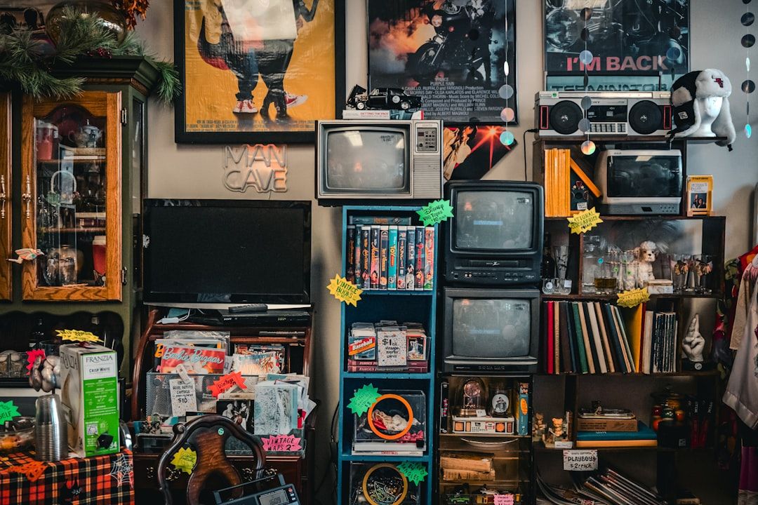Ein überfüllter Raum mit verschiedenen Vintage-Elektronikgeräten, Büchern, Schallplatten, Postern und Erinnerungsstücken. Unter den Gegenständen sind mehrere Röhrenfernseher und ein Aachen-Reiseposter ausgestellt, zusammen mit einem „Man Cave“-Schild.