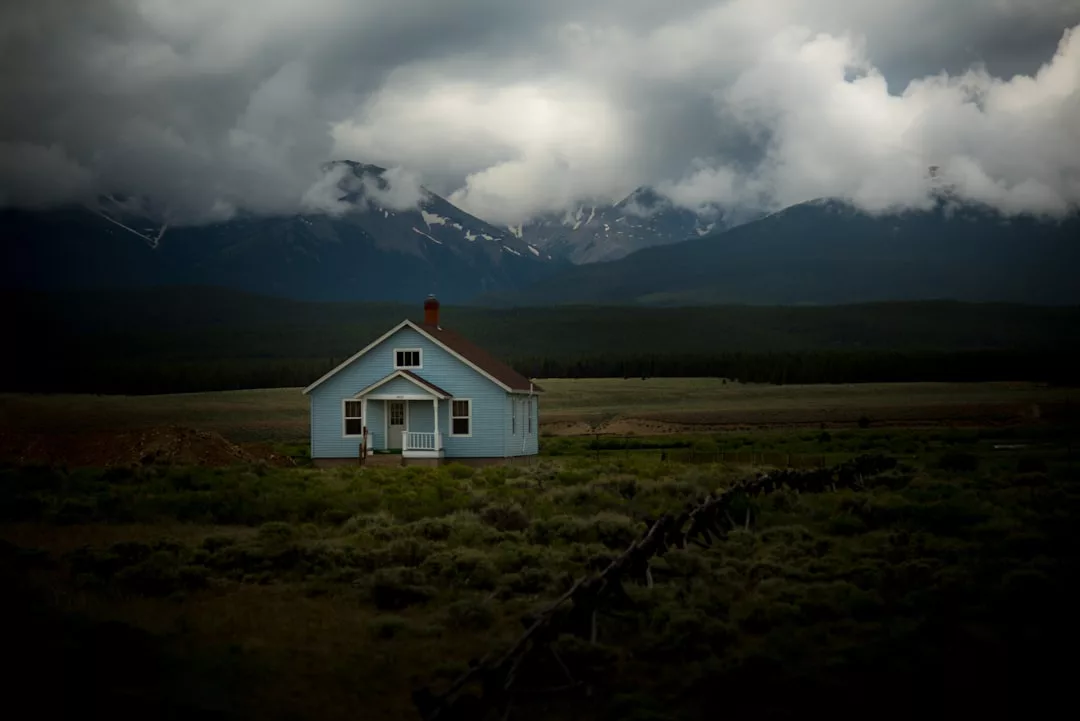 Ein kleines blaues Haus steht isoliert auf einem riesigen Feld, umgeben von Bergen und dunklem, bewölktem Himmel.
