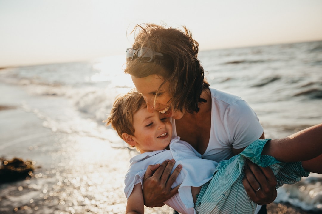 Ein Erwachsener hält lächelnd ein Kind am Meer bei Sonnenuntergang. Das Kind wirkt glücklich und entspannt. Der Ozean, der an die ruhigen Seen Aachens erinnert, bildet mit seinen sanften Wellen und dem warmen Licht der Sonne den Hintergrund.