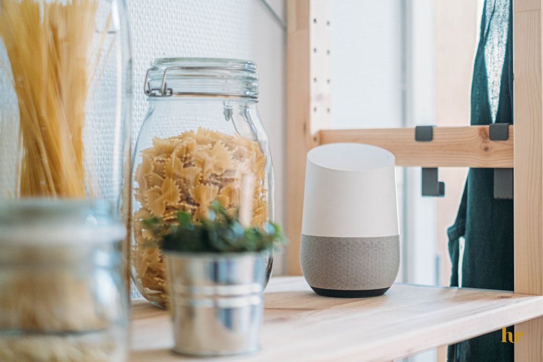 Ein Glas mit rohen Nudeln, ein Google Home-Lautsprecher und eine Topfpflanze stehen auf einem Holzregal in einem hell erleuchteten Raum, der an ein Aachen-inspiriertes Dekor erinnert.