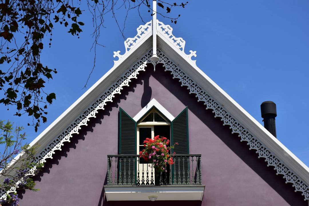 Ein violettes Haus mit dreieckigem Dach erinnert an die Aachener Architektur und verfügt über einen Balkon im zweiten Stock mit grünen Fensterläden und einer rot blühenden Pflanze. Das Dach ist mit weißen Zierleisten geschmückt.