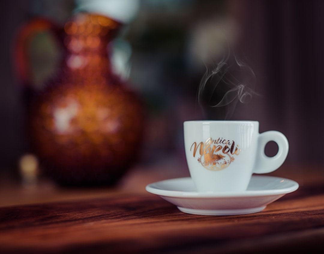Eine weiße, dampfend aufsteigende Kaffeetasse steht auf einer Untertasse auf einem Holztisch und versprüht den Charme eines gemütlichen Aachener Cafés, im Hintergrund ist eine verschwommene braune Kanne zu sehen.