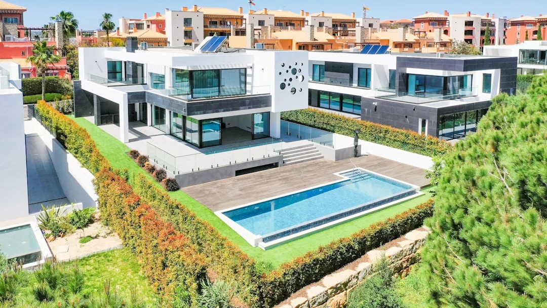 Das moderne, weiß-schwarze Haus mit großen Fenstern ist von angelegten Grünflächen umgeben, verfügt über einen rechteckigen Swimmingpool und liegt in einem Wohngebiet mit Häusern im ähnlichen Stil im Hintergrund.