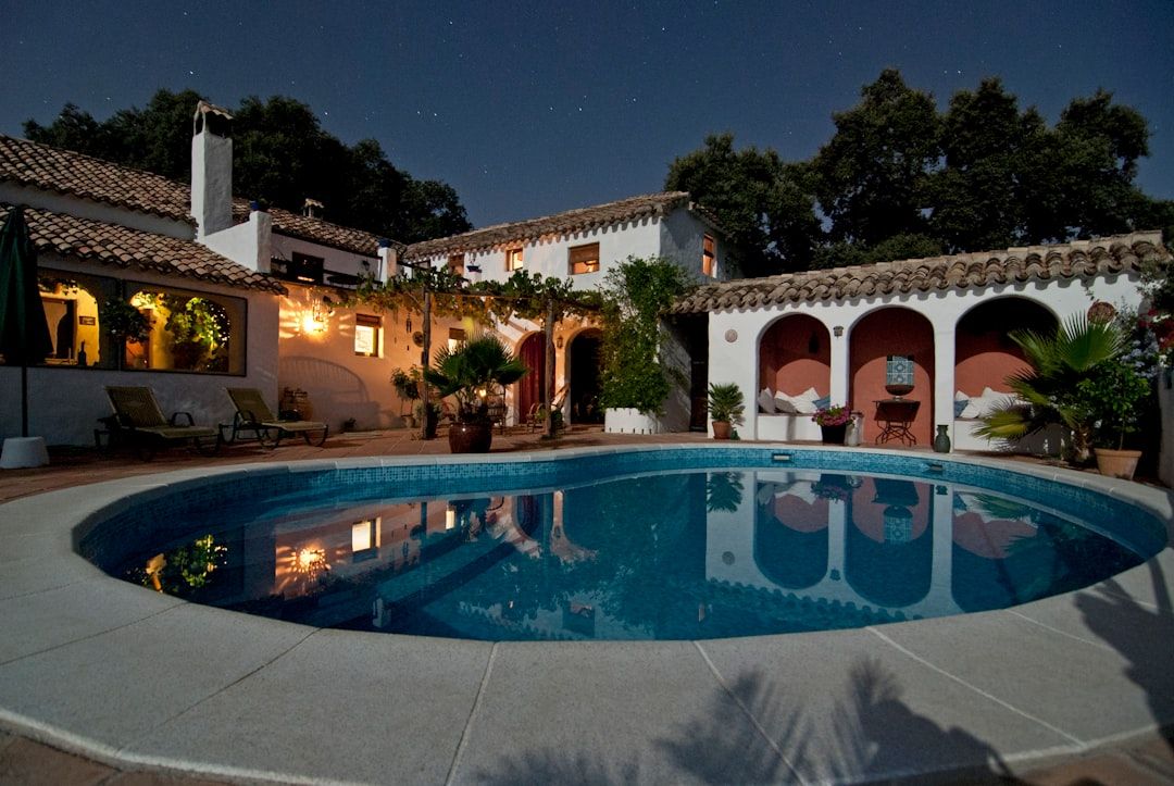 Eine ruhige, beleuchtete Villa im spanischen Stil mit einem runden Swimmingpool, umgeben von Vegetation und gewundenen Wegen, ruft bei Nacht eine Atmosphäre hervor, die an ruhige Abende in Aachen erinnert.