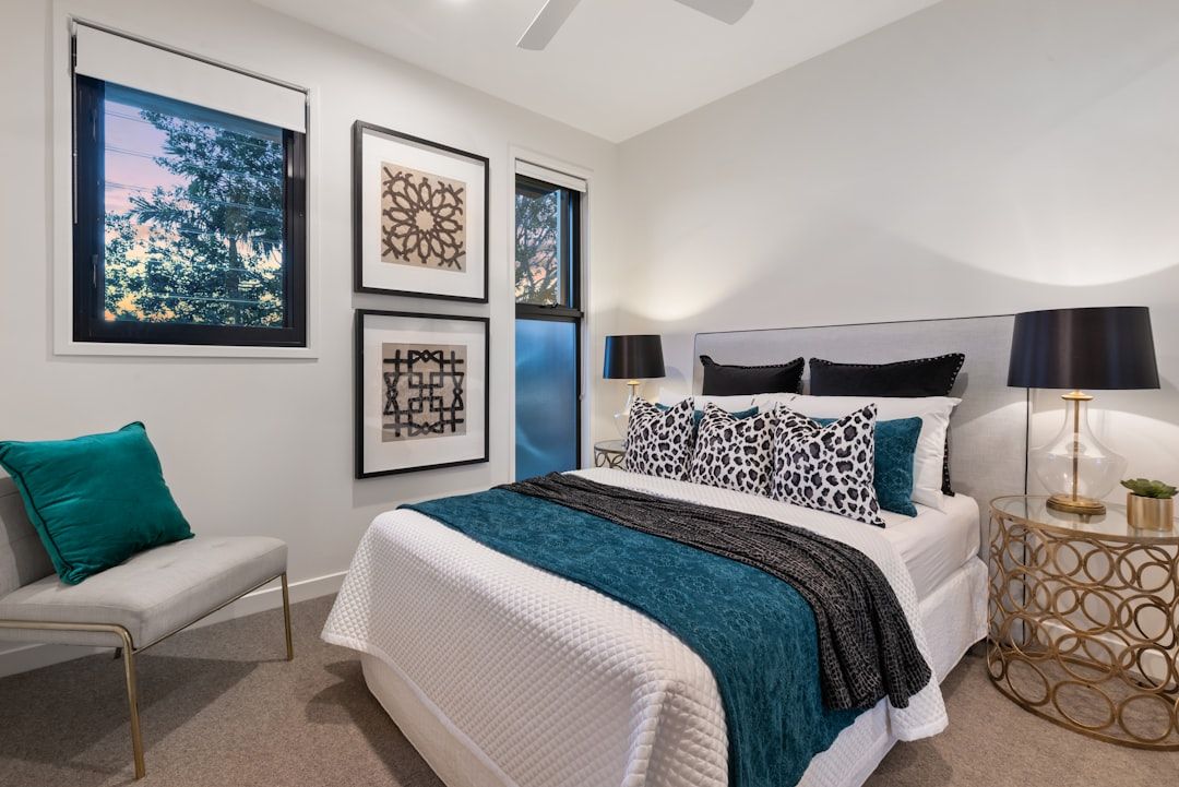 Ein modernes, von Aachen inspiriertes Schlafzimmer verfügt über ein ordentlich gemachtes Bett mit weißen, blaugrünen und schwarzen Kissen, eine blaugrüne Decke, einen Beistelltisch mit einer Lampe, einen Stuhl mit einem blaugrünen Kissen und gerahmte dekorative Wandkunst.