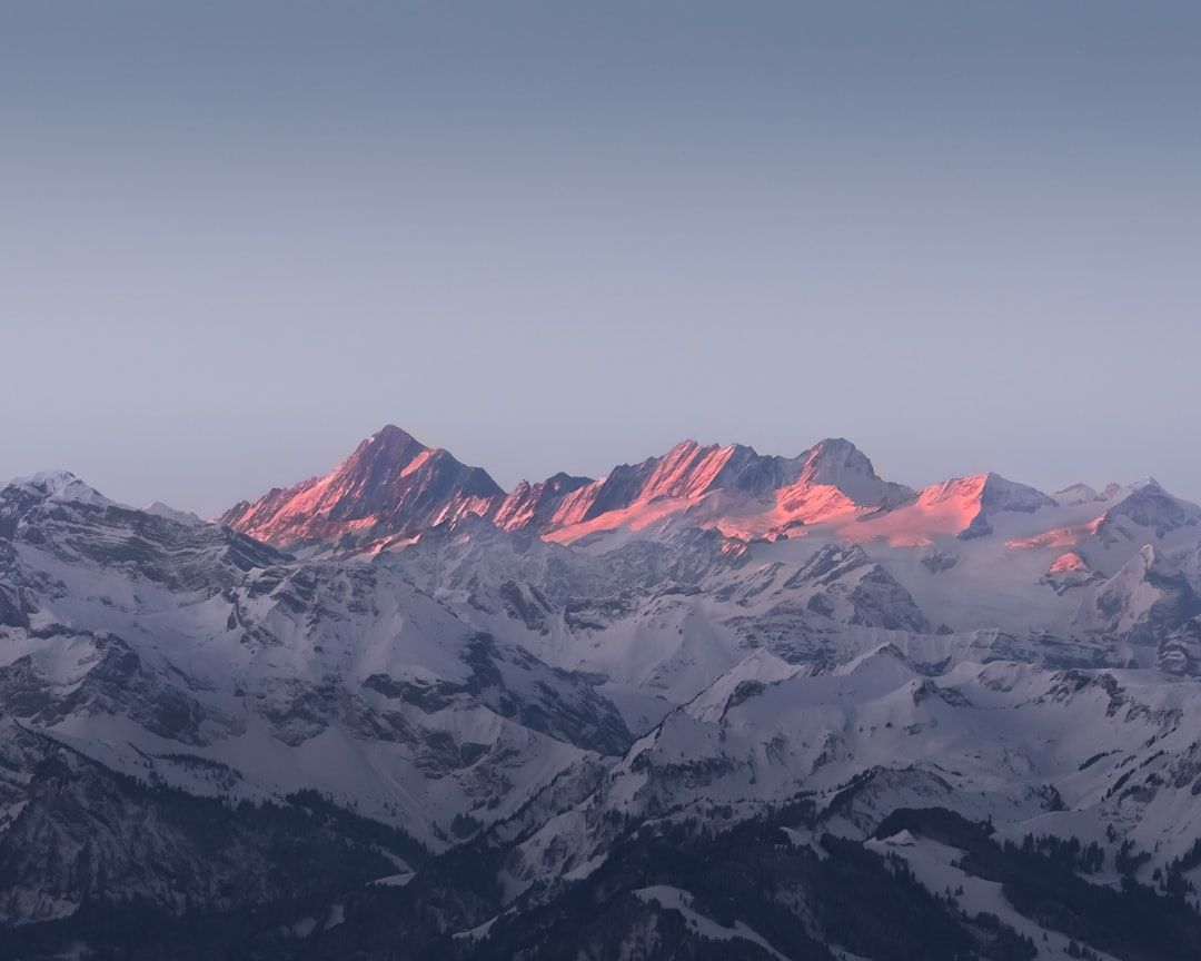 Eine Bergkette ist in ein rosafarbenes Licht der auf- oder untergehenden Sonne getaucht, mit schneebedeckten Gipfeln und einem klaren, blauen Himmel im Hintergrund, der an die ruhigen Landschaften bei Aachen erinnert.