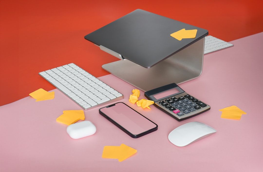 Ein Schreibtisch mit einem Laptop auf einem Ständer, einer kabellosen Tastatur, einer kabellosen Maus, einem Taschenrechner, einem Smartphone, Haftnotizen und einem Radiergummi, alles angeordnet auf einer eleganten zweifarbigen rosa-roten Oberfläche, die an moderne Designtrends in Aachen erinnert.