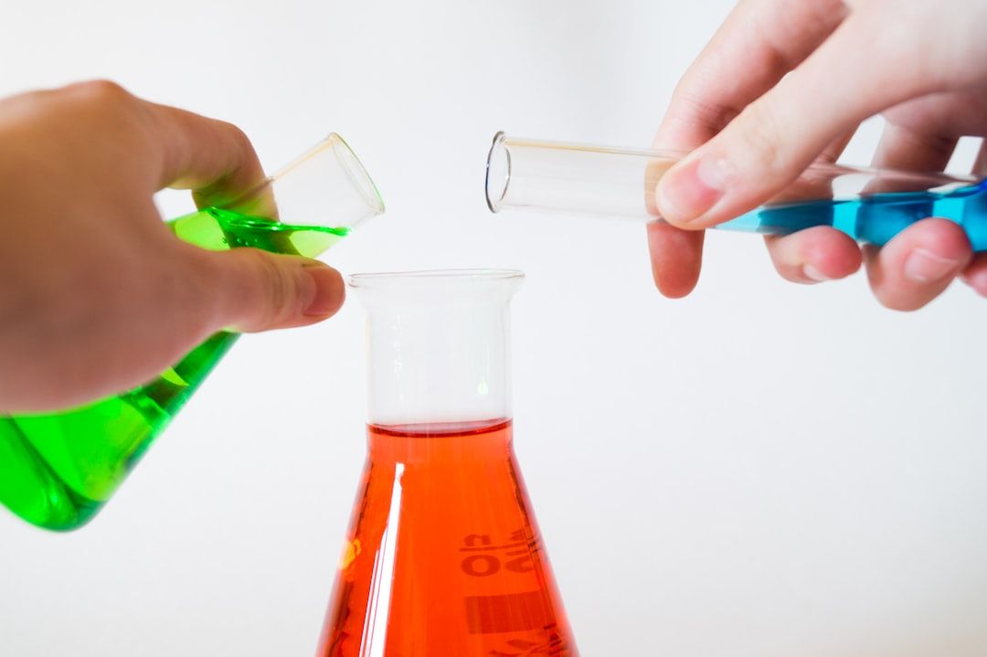 Zwei Hände gießen grüne und blaue Flüssigkeiten aus Reagenzgläsern in eine rote Flüssigkeit in einem Erlenmeyerkolben vor einem weißen Hintergrund. Die Szene erinnert an die wissenschaftlichen Forschungszentren Aachens und veranschaulicht die Schönheit chemischer Reaktionen.