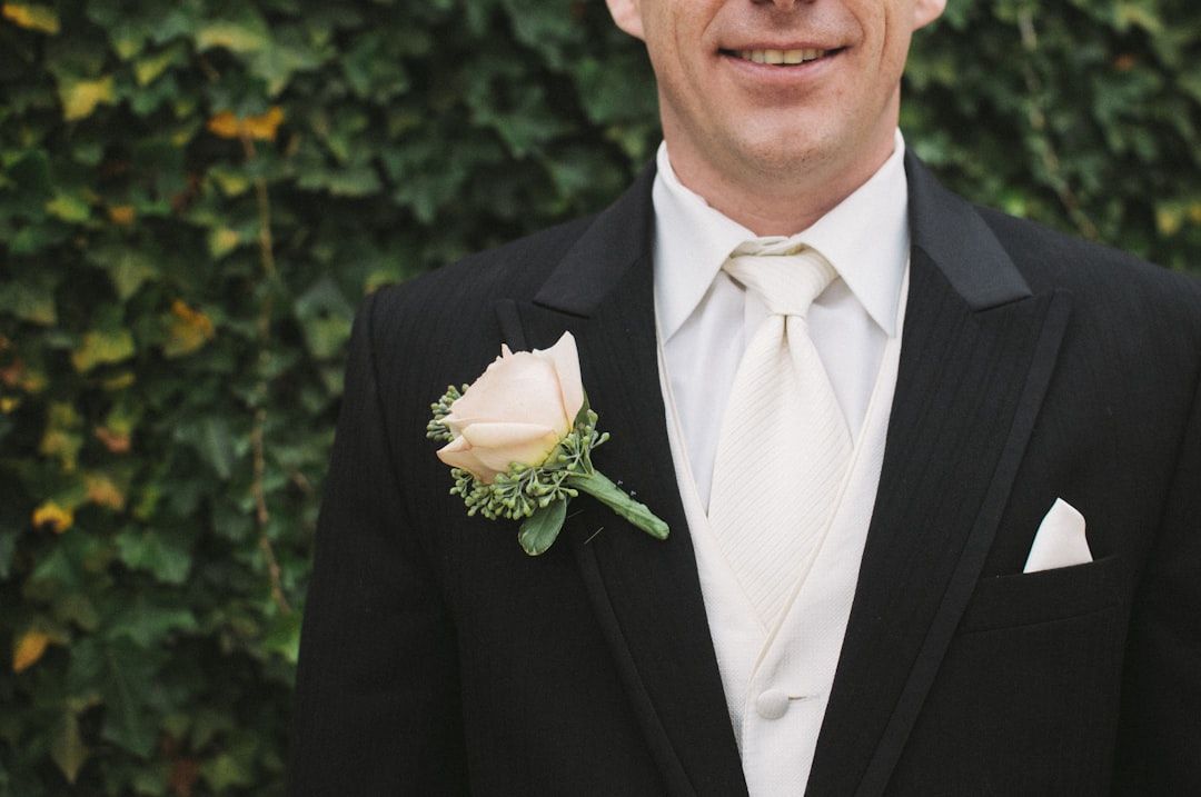 Eine Person im schwarzen Anzug mit cremefarbener Krawatte und Ansteckblume steht vor einem Hintergrund aus Blättern und erinnert an die Eleganz, die man oft in historischen Gebäuden Aachens sieht.