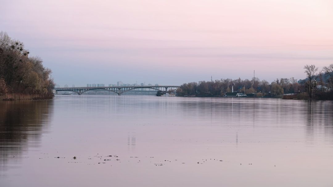 Ein breiter, ruhiger Fluss, der an die heitere Landschaft Aachens erinnert, mit einer in der Ferne gespannten Brücke, gesäumt von Bäumen mit spärlichem Laub unter einem frühen Morgenhimmel.
