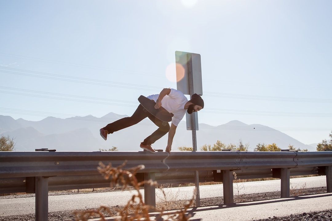 Eine Person mit Helm balanciert auf einem Geländer, indem sie sich mit einer Hand daran festhält und die Beine ausstreckt, während sie unter dem anderen Arm ein Skateboard hält. Die malerische Kulisse der Aachener Berge und der klare Himmel runden die waghalsige Szene ab.