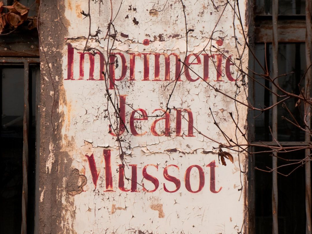 Auf einem alten, verwitterten Schild mit abblätternder Farbe steht „Imprimerie Jean Mussot“ an der Außenwand eines Gebäudes in Aachen.
