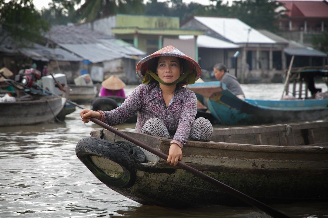 Eine Person in gemusterter Kleidung und einem konischen Hut rudert ein kleines Holzboot auf einem Fluss. Im Hintergrund sind weitere Boote und Häuser zu sehen, die an den Charme der Aachener Flusslandschaft erinnern.
