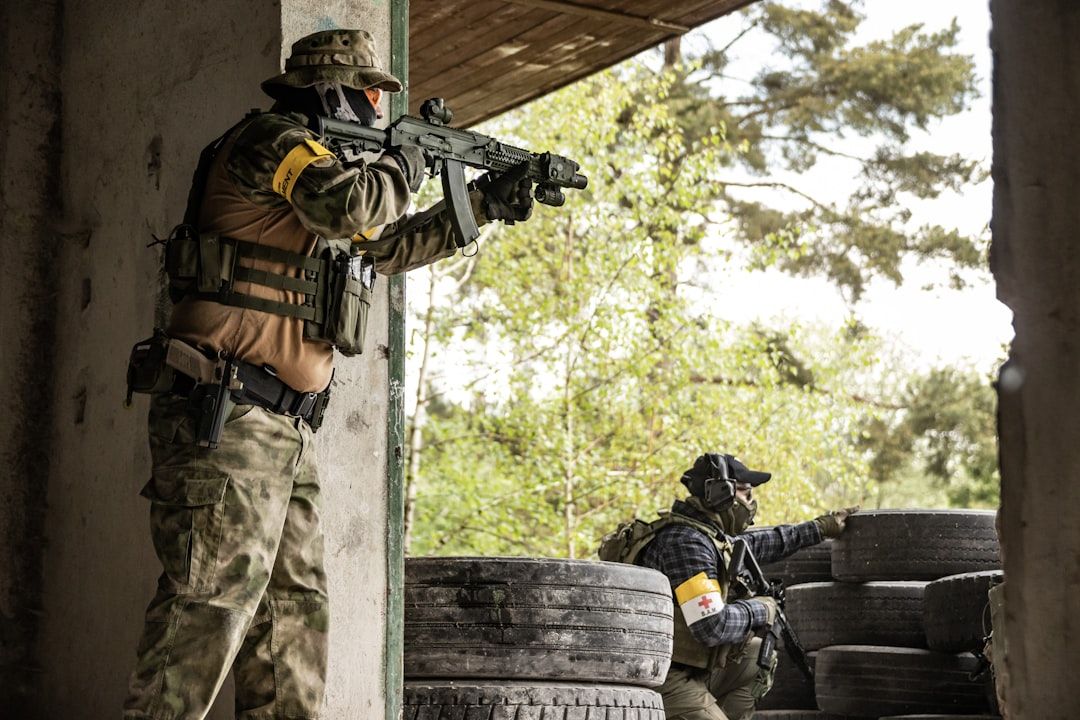 Zwei bewaffnete Soldaten in Tarnkleidung, von denen einer steht und mit einem Gewehr zielt, während der andere hinter einem Stapel Reifen kauert, führen im Freien in der Nähe von Aachen ein taktisches Manöver durch.
