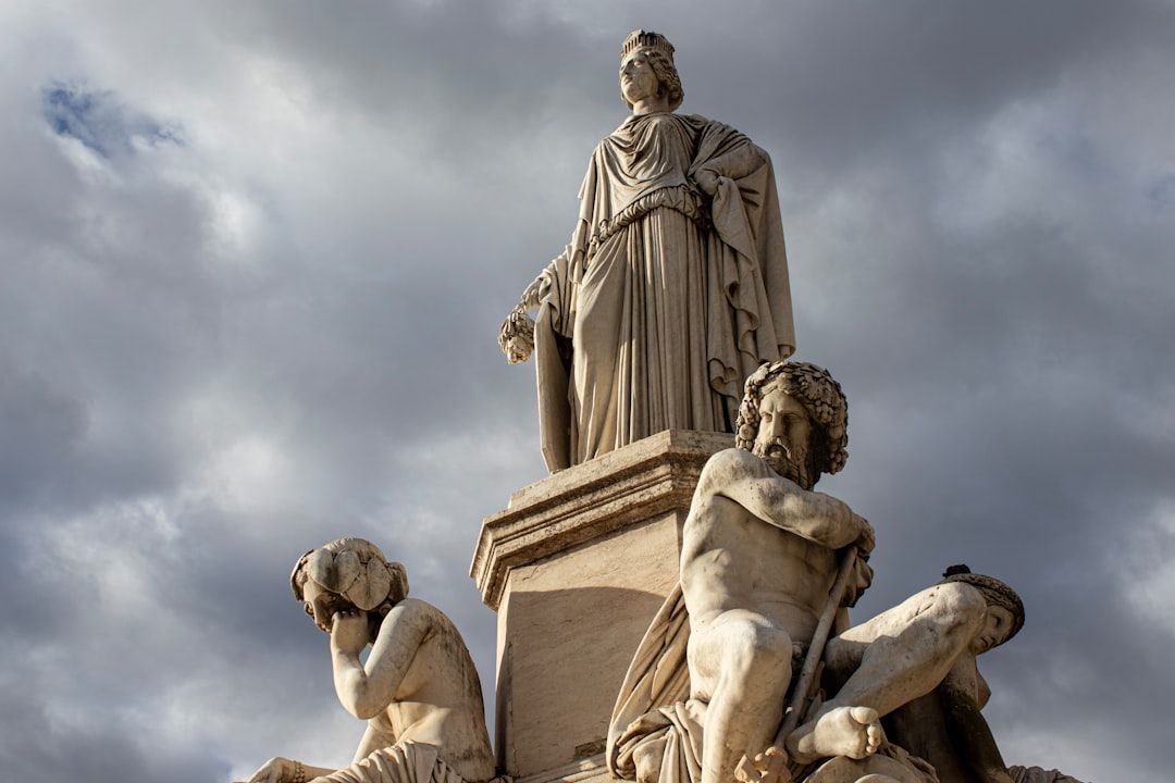 Statue mit drei Figuren: einer stehenden zentralen Figur im Gewand und zwei sitzenden Figuren auf beiden Seiten, vor dem wolkigen Himmel von Aachen.