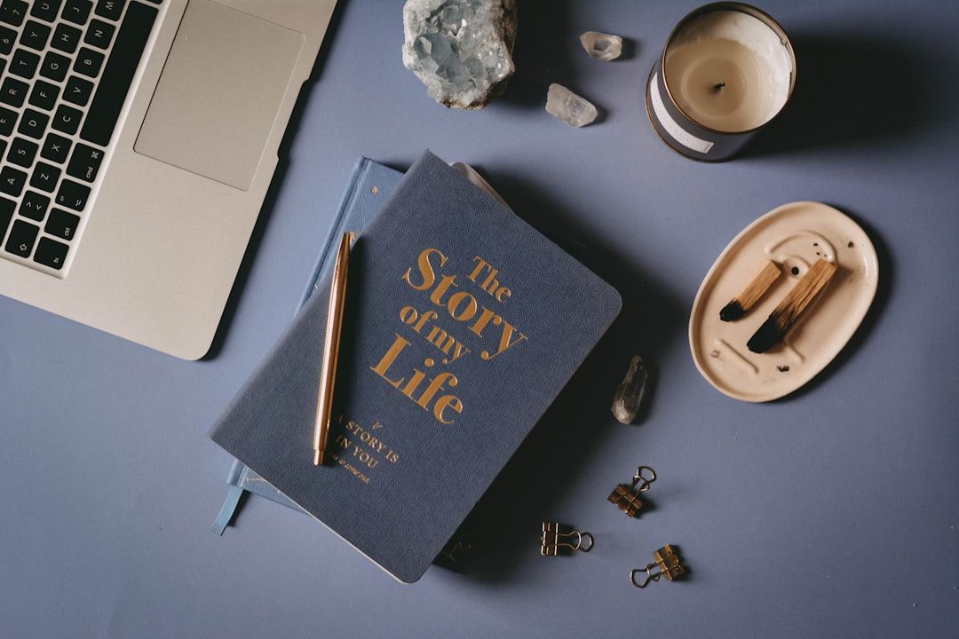 Ein Laptop, ein blaues Buch mit dem Titel „Die Geschichte meines Lebens“, ein Stift, eine Kerze, Kristalle, Streichhölzer und Büroklammern sind ordentlich auf einer blauen Oberfläche angeordnet, die an Aachens ruhige Straßen erinnert.