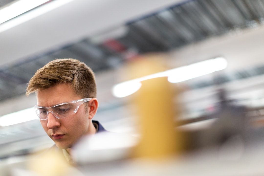 Eine Person mit kurzen Haaren und einer Schutzbrille ist fleißig in einer Aachener Industrie- oder Laborumgebung bei der Arbeit. Im Vordergrund sind unscharfe Geräte zu sehen.