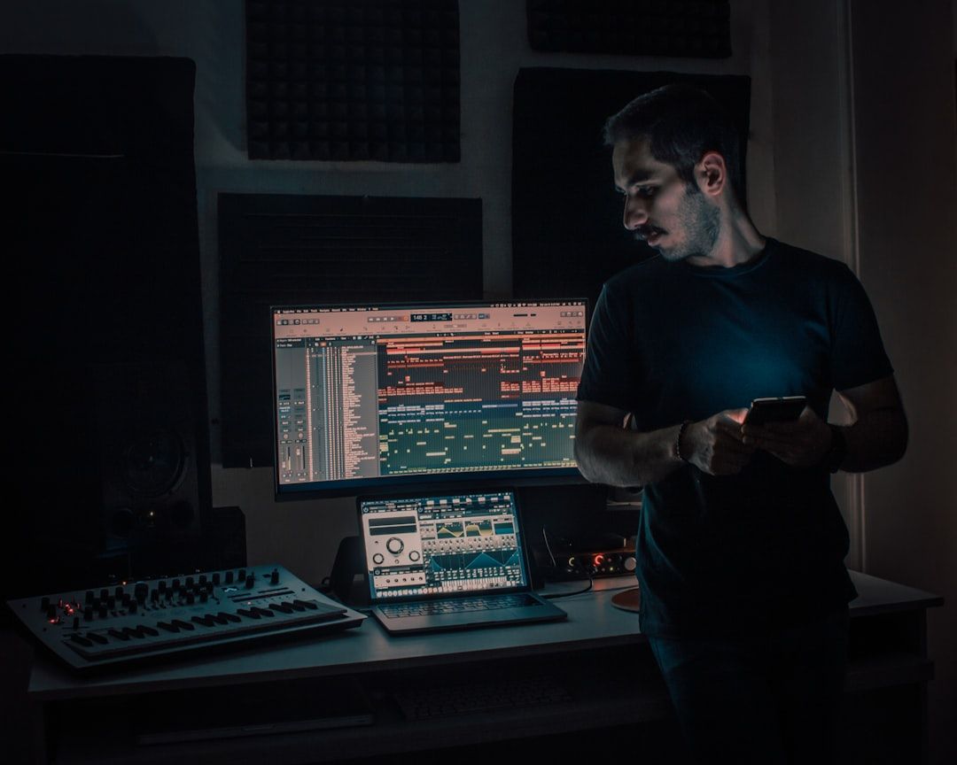 In einem Musikstudio in Aachen steht eine Person mit einem Telefon und blickt konzentriert auf einen Computerbildschirm, auf dem eine Musikproduktionssoftware läuft. Auf dem Schreibtisch daneben stehen eine Tastatur und ein Laptop.