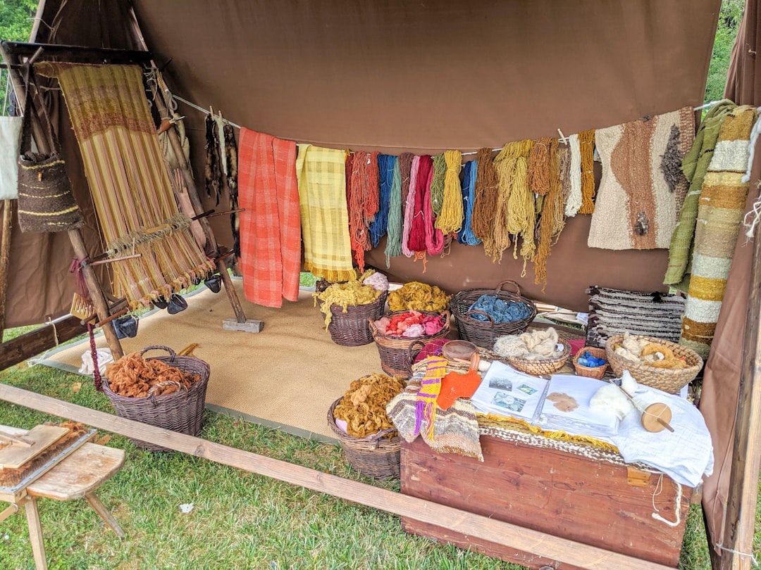 Ein Stand auf einem Freiluftmarkt im Herzen von Aachen bietet gewebte Textilien, Garne und Körbe mit Fasern, die auf Tischen und hängenden Displays angeordnet sind. Garne in verschiedenen Farben werden prominent präsentiert und bilden ein lebendiges Bild, das den Blick auf sich zieht und zum Erkunden einlädt.