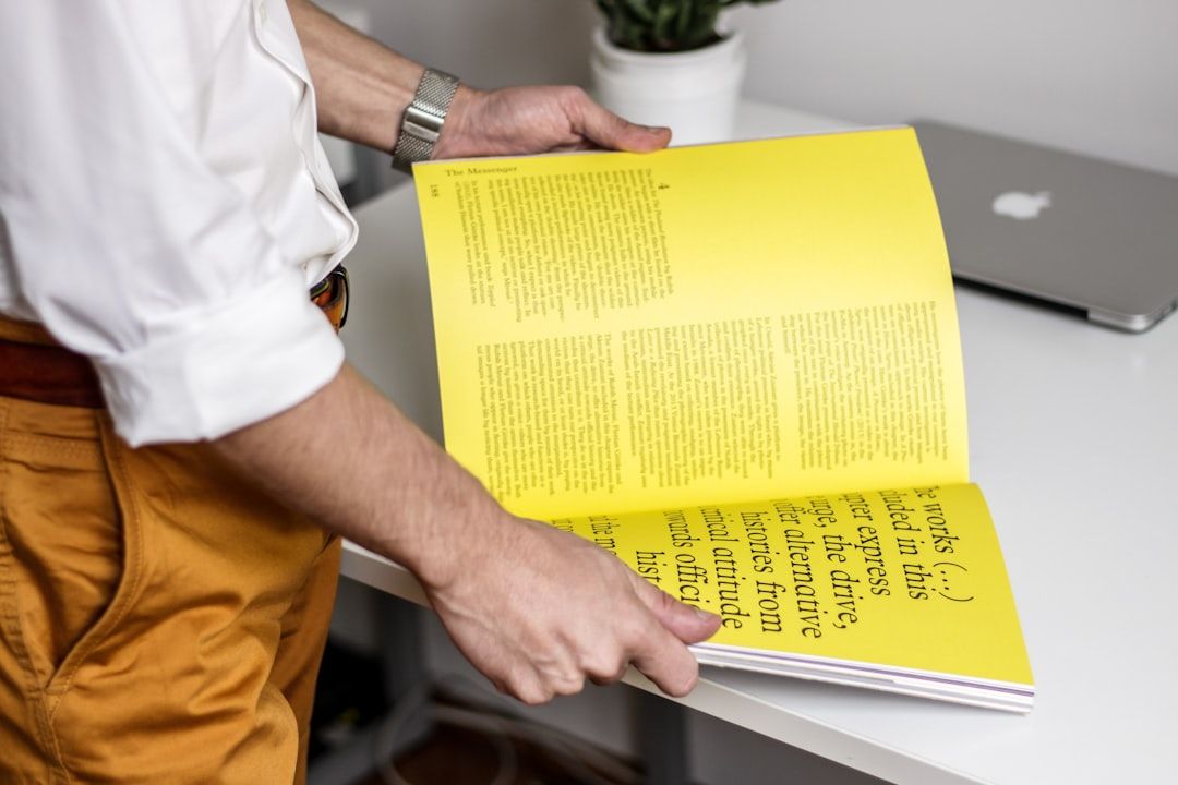 Eine Person in weißem Hemd und gelber Hose hält eine gelbe Zeitschrift mit Text aufgeschlagen in der Hand, steht neben einem Schreibtisch mit Laptop und einer kleinen Topfpflanze und träumt vielleicht von ihrer nächsten Reise nach Aachen.