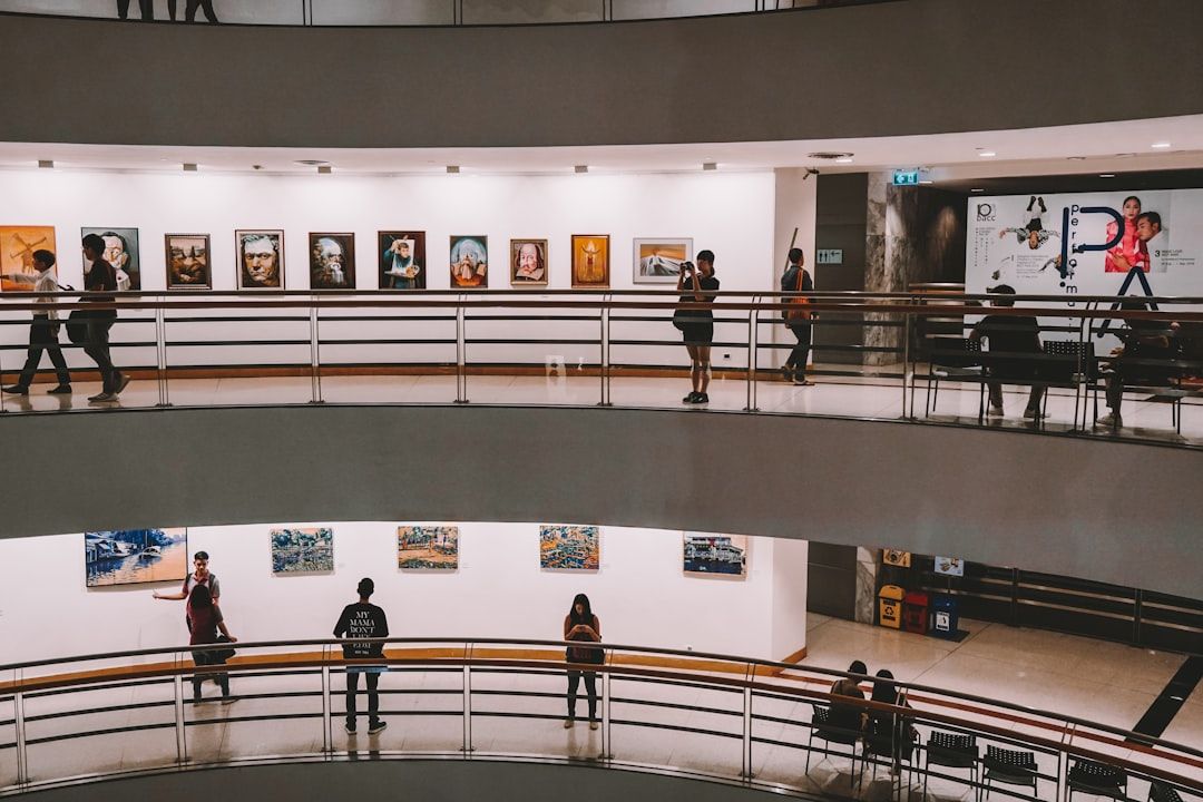 Zwei Ebenen einer Kunstgalerie, in der Menschen die an den Wänden hängenden Gemälde und Fotografien betrachten. Die von der Eleganz Aachens inspirierte Galerie ist gut beleuchtet und modern mit einem eleganten Geländerdesign.