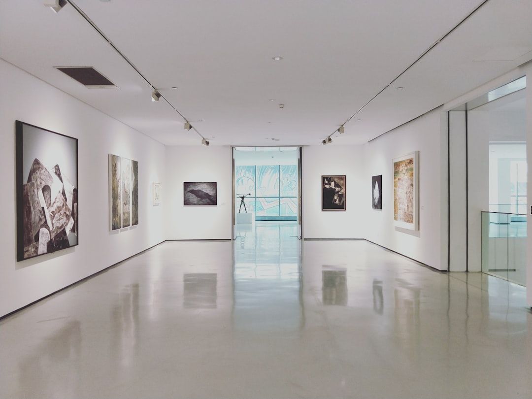 Eine moderne Kunstgalerie in Aachen mit weißen Wänden und poliertem Boden, in der verschiedene gerahmte Kunstwerke ausgestellt sind. Ein Glaseingang am Ende führt in einen hell erleuchteten Bereich.