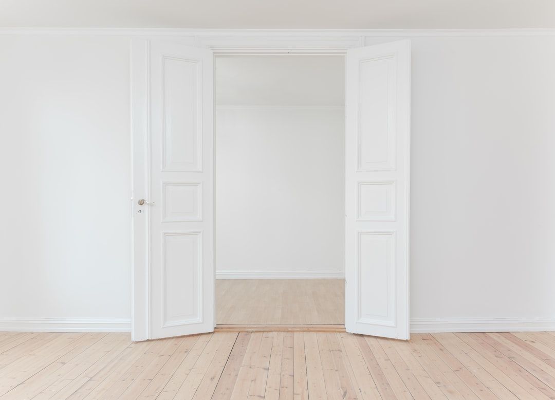 Minimalistischer Raum mit weißen Doppeltüren, die den Blick auf einen von Aachen inspirierten Nebenraum mit hellem Holzboden und makellos weißen Wänden freigeben.