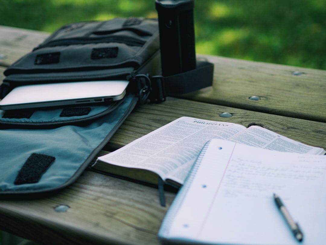 Offene Laptoptasche, Notizbuch und Stift auf einem hölzernen Picknicktisch in Aachen. Aufgeschlagene Bibelseite neben dem Notizbuch. Außenkulisse mit grünem, verschwommenem Hintergrund.