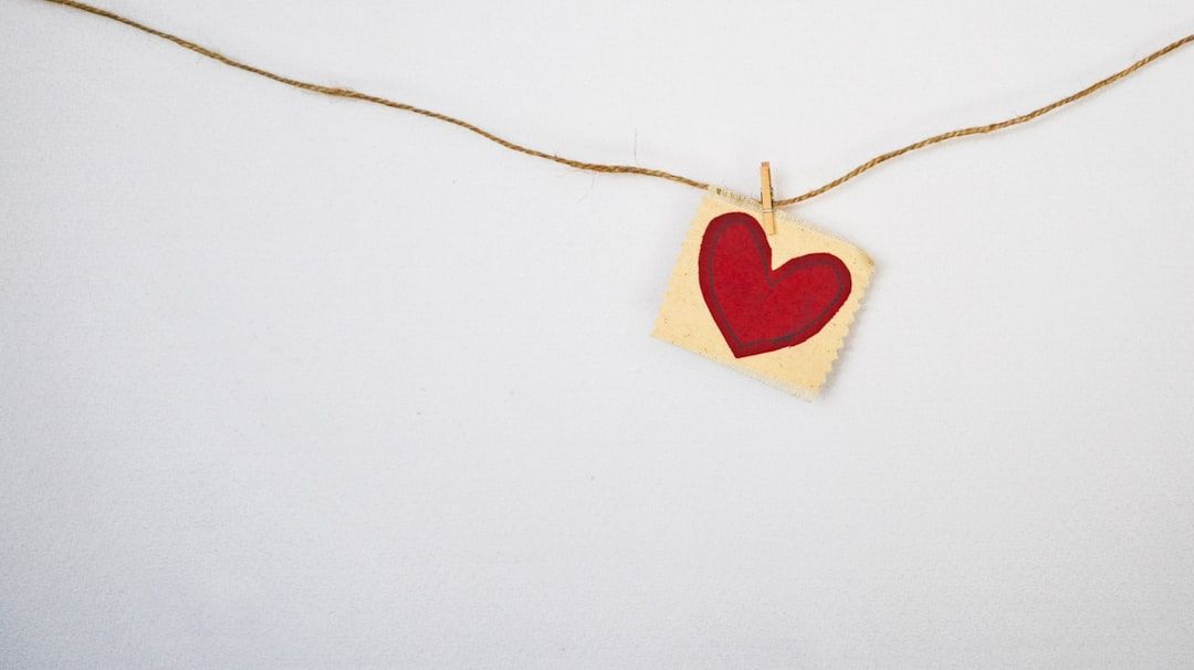 Ein rotes Herz auf einem kleinen Stück Papier ist an einer braunen Schnur befestigt und erinnert an eine Kunstinstallation, die man in den charmanten Straßen von Aachen finden würde, und das alles vor einem schlichten weißen Hintergrund.