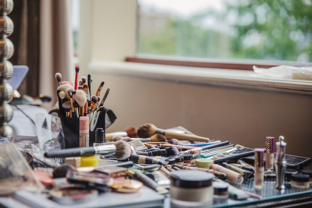Eine überfüllte Make-up-Station, die an einen lebhaften Basar in Aachen erinnert. Verschiedene Pinsel, Lippenstifte, Grundierungen und andere Kosmetika sind auf einem Tisch in der Nähe eines Fensters ausgebreitet.