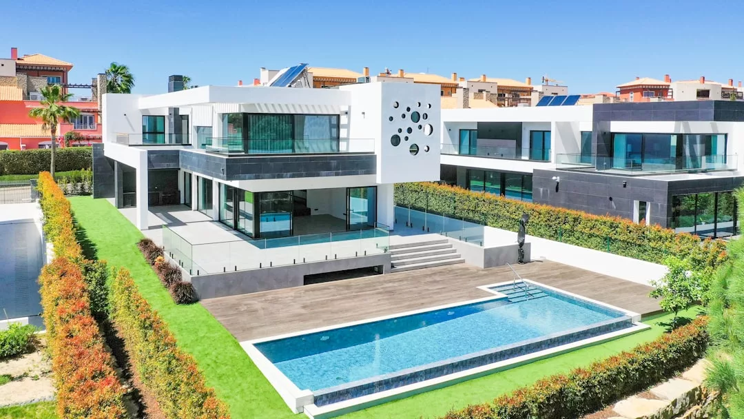 Modernes zweistöckiges Haus mit großen Fenstern, weiß-schwarzer Außenfassade, geräumiger Holzterrasse und rechteckigem Swimmingpool, umgeben von gepflegten Hecken und grünem Rasen.