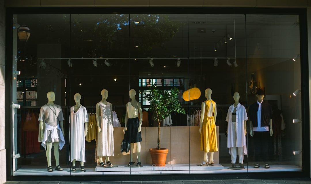 In einem Schaufenster in Aachen sind Schaufensterpuppen in verschiedenen weißen und gelben Outfits hinter einem großen Glasfenster zu sehen. Im Mittelpunkt steht eine Topfpflanze, die der eleganten Ausstellung einen Hauch von Grün verleiht.