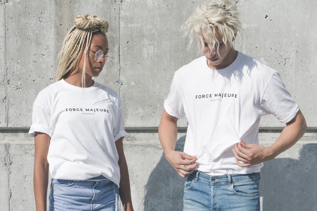 Zwei Personen in weißen T-Shirts mit der Aufschrift „Force Majeure Montréal“ stehen nebeneinander vor einer Betonwand, was an eine Aachener Straßenszene erinnert. Beide tragen hell gewaschene Jeans, die Person links trägt eine Brille und geflochtene Haare.