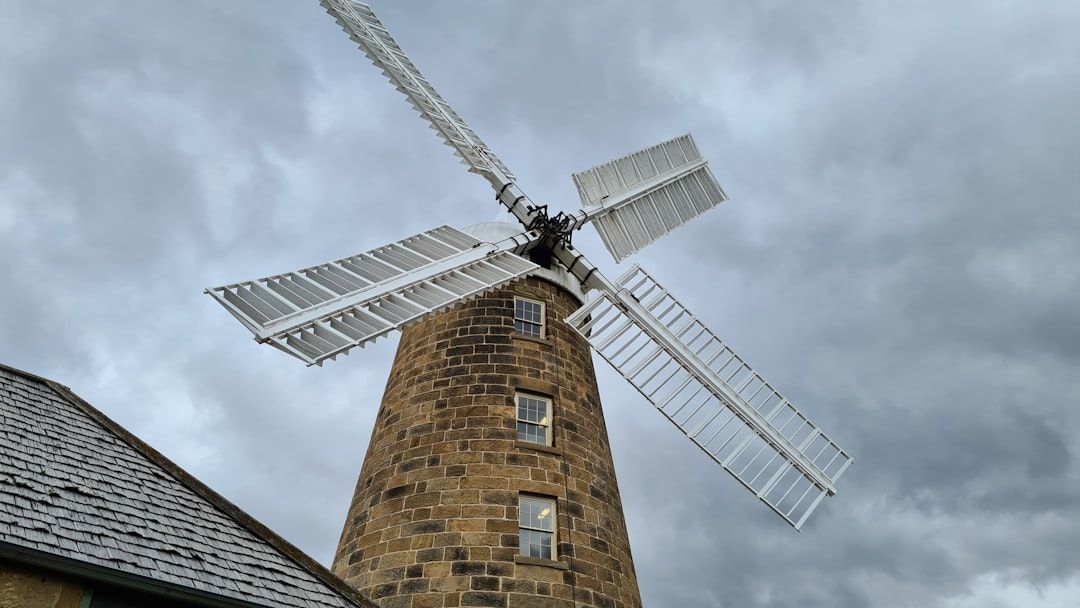 Vor einem wolkigen Himmel in Aachen steht eine steinerne Windmühle mit weißen Flügeln, begleitet von einem Gebäudeteil mit Schindeldach.