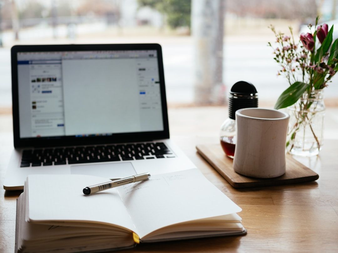 Ein Laptop, ein aufgeschlagenes Notizbuch mit Stift, eine Tasse Kaffee und eine kleine Vase mit Blumen stehen auf einem Holztisch am Fenster und vermitteln die gemütliche Atmosphäre eines Aachener Cafés.