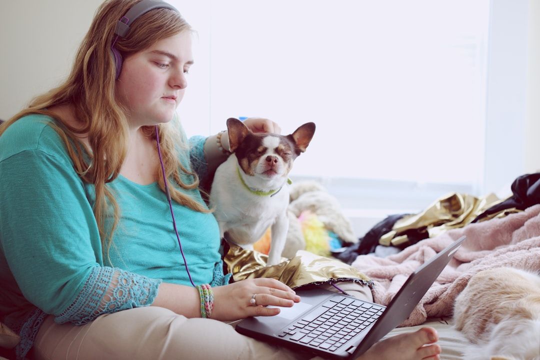 Eine Person sitzt auf einem Bett in einer Aachener Wohnung, arbeitet mit Kopfhörern an einem Laptop und streichelt einen kleinen Hund.