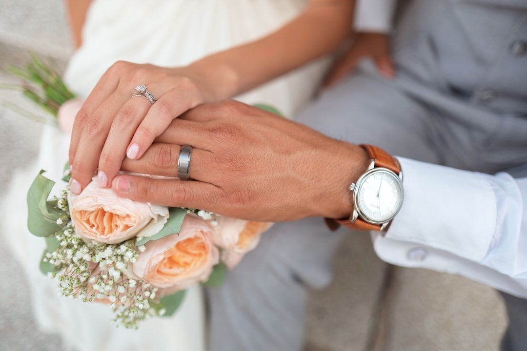 Zwei mit Eheringen geschmückte Hände ruhen auf einem Strauß aus Pfirsichrosen und Schleierkraut in Aachen. An einem Zeiger hängt ein Diamant-Verlobungsring, am anderen eine silberne Uhr. Beide tragen elegante Hochzeitskleidung.